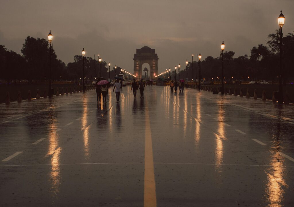 Moody evening scene at India Gate with reflections on the wet road and people walking.