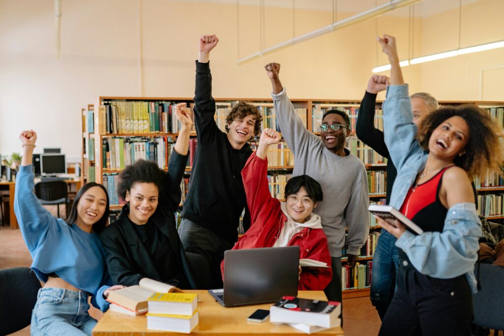 Happy diverse students in library raising hands, enjoying academic success.